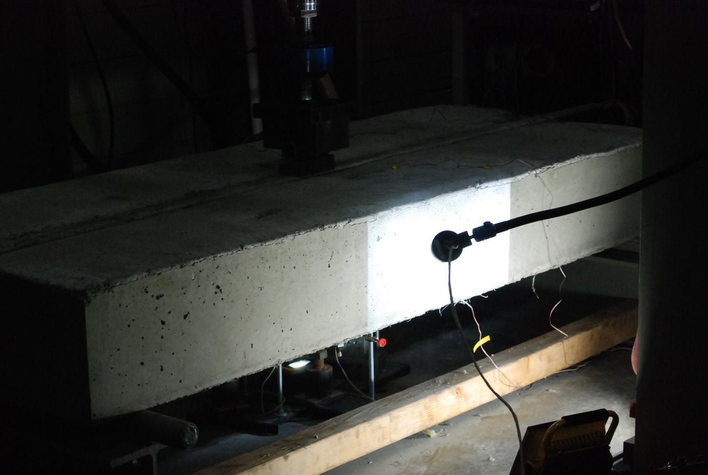 A concrete beam under a loading head in a test frame, lit from the side